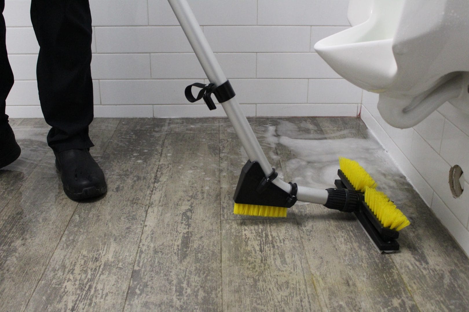 Person scrubbing a restroom floor with a yellow-bristled cleaning brush near a urinal